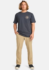 Mørkegrå bomulls T-skjorte med et sirkulært logo på brystet, parret med lyse beige bukser og svarte joggesko. Stående mannlig modell.