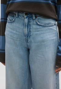 Ανοιχτό μπλε τζιν τζιν με χαλαρή εφαρμογή, που διαθέτει μπροστινές τσέπες, κλείσιμο με κουμπί και αντίθετη ραφή. Συνδυασμένο με σκούρο ριγέ τοπ.