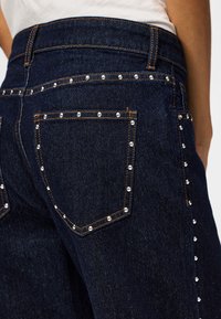 Jeans en denim bleu foncé avec des clous en argent dessinant la poche arrière et la couture latérale, portés avec un haut blanc.