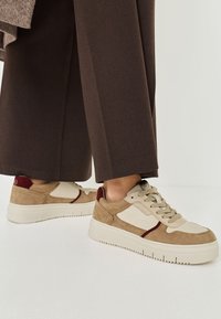 Beiga och burgundy sneakers med mocka och strukturerade paneler, med en tjock vit sula och strukturerade snören, placerade mot bruna byxor.