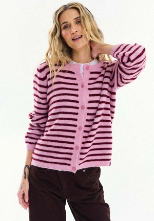 Kobieta o falowanych blond włosach nosi różowo-bordowy sweter na guziki w paski oraz ciemne spodnie, pozuje z jedną ręką założoną na kark.