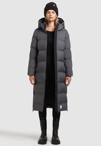 Lange, puffy, donkergrijze donsjas met een capuchon, voorzien van een ritssluiting, zij-ritsen en een logo-tag aan de zoom. Draagt bij zwarte leggings en laarzen.