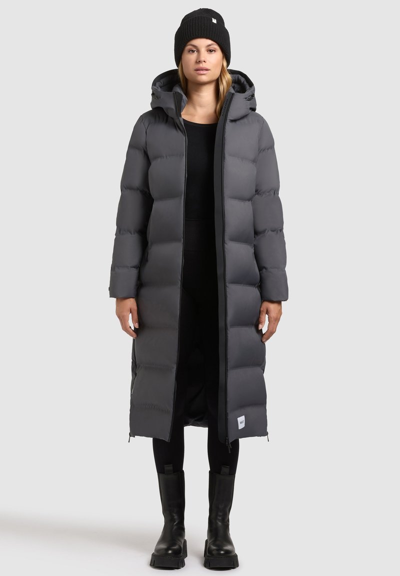 Lange, puffy, donkergrijze donsjas met een capuchon, voorzien van een ritssluiting, zij-ritsen en een logo-tag aan de zoom. Draagt bij zwarte leggings en laarzen.