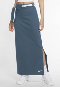 Jupe Nike bleu foncé, fabriquée en tissu doux. Elle présente une fente latérale, des accents blancs avec une ceinture, et un logo sur l'ourlet. Portée avec des baskets blanches.