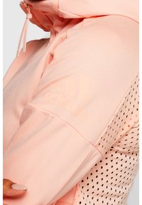 Zbliżenie na osobę noszącą jasnoróżową bluzę Adidas z kapturem, zamkiem błyskawicznym, sznurkiem oraz perforowanymi detalami wentylacyjnymi na rękawie.