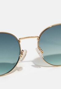 Gafas de sol con montura metálica dorada y grandes lentes redondos con degradado, que van de un azul oscuro en la parte superior a un tono claro en la parte inferior. Almohadillas para la nariz transparentes adjuntas.