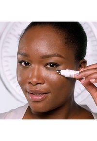 White beauty applicator with a silver tip held near the eye of a woman with dark skin. Background features a circular clock design.