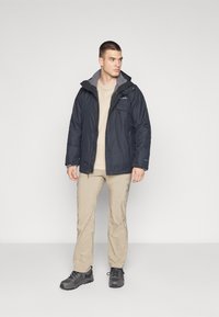 Dark navy insulated jacket with a gray fleece lining, front zipper, and two chest pockets, worn over a light beige shirt and khaki pants.