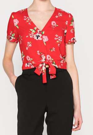 Rød kortærmet bluse med blomstermønster, frontknapper og bundet talje, båret med højtaljede sorte bukser, hånd i lommen.