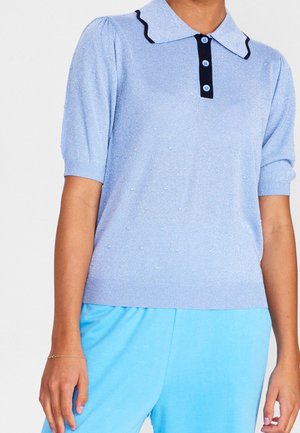 Femme portant un haut tricoté à manches courtes bleu clair avec un col festonné et une patte de boutonnage noire, associé à un pantalon bleu vif.