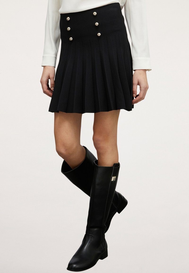 Femme portant une jupe plissée noire avec des boutons décoratifs, un haut blanc à manches longues et des bottes en cuir noires montantes jusqu'aux genoux.
