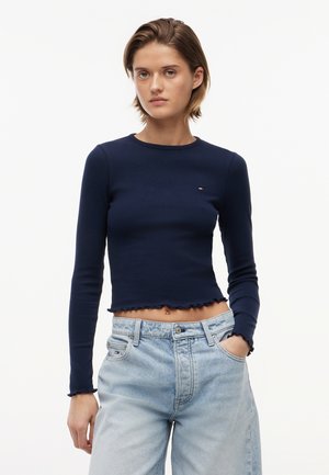 Kobieta w granatowym, prążkowanym topie z długim rękawem o falowanych krawędziach oraz jasnoniebieskich, wysokich dżinsach z rękami w kieszeniach.