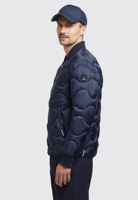 Mörkblå puffjacka med ett kviltat vågmönster, lättviktig glänsande tyg, ribbad krage, dragkedjefickor på sidan och logotypdetalj på vänster ärm.
