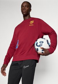 Μαρόν φούτερ με λογότυπο της Μπαρτσελόνα, διακοσμητική πλαϊνή ραφή, σε συνδυασμό με μαύρο παντελόνι Nike. Το μοντέλο κρατάει μια λευκή και μπλε ποδοσφαιρική μπάλα.