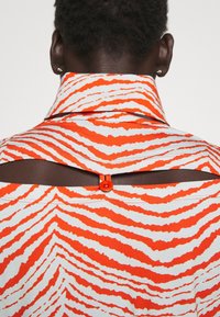 Skjorta med ett orange och vitt zigzag-mönster, som har en hög krage, en öppning på ryggen och knäppning vid halsen. Tillverkad av lättviktig tyg.