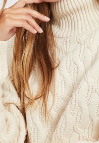 Pull col montant en tricot épais de couleur crème, avec un col côtelé et un motif de tricot en câble, affichant une texture douce et une coupe ample.