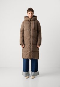 Manteau en duvet marron avec capuche, doté d'une fermeture éclair, de cordons de serrage réglables et d'un design en coutures horizontales ; associé à un pantalon en denim à jambes larges.