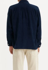 Marinblå kordflanellskjorta med knappkrage, långa ärmar och veckad detalj bak. Stylingen inkluderar ljusa, lediga byxor.