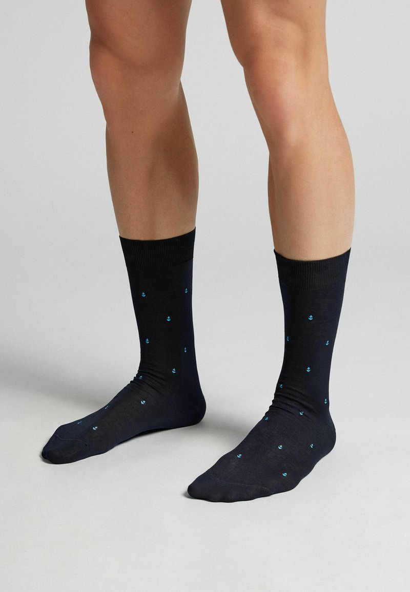 Jambes portant des chaussettes noires mi-mollet avec de petits motifs d'ancre bleue, debout sur une surface gris clair.