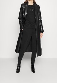 Lång svart läderjacka över en svart sweatshirt med paljettdetaljer, ihopparat med svarta byxor och ankelboots, vilket skapar en monokromatisk look.