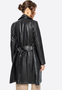 Abrigo de cuero negro con una silueta entallada, mangas largas y un lazo en la cintura. Textura suave con un cuello clásico y detalles en la costura trasera.
