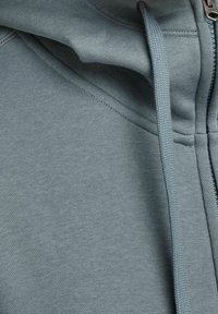 Felpa con zip in tessuto verde teal tenue, caratterizzata da una superficie liscia, cappuccio con cordino e dettagli di cucitura visibili lungo i bordi.