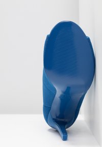 Chaussure à talons bleue positionnée la semelle vers le haut contre un coin blanc, montrant le dessous lisse et texturé ainsi que le talon étroit.