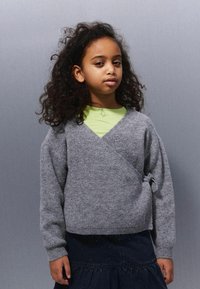 Djevojka s kovrčavom tamnom kosom nosi sivi džemper s preklopom preko svijetlozelenog majice i tamne suknje, stojeći protiv jednostavne sivoj pozadini.