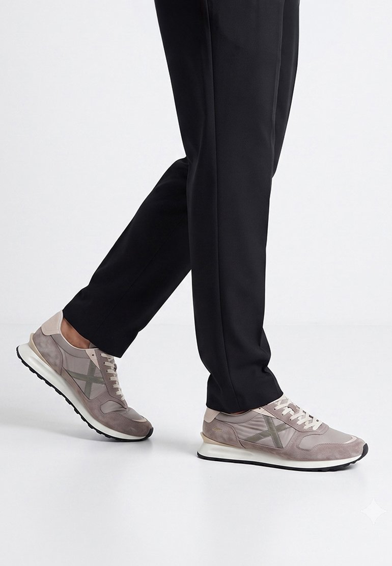 Personne portant un pantalon noir et des baskets décontractées gris clair debout sur un sol blanc, un pied légèrement levé.