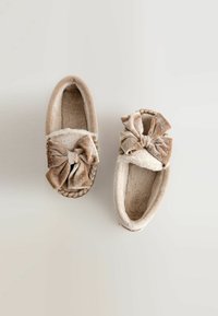 Chaussons beiges en tissu doux avec une doublure en peluche. Chacun présente un grand nœud en tissu, l'un d'eux ayant un motif à pois.