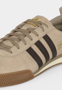 Baskets Adidas en daim beige avec trois bandes marron, lacets beige, semelle en gomme naturelle et logo Adidas doré sur la languette et le côté.