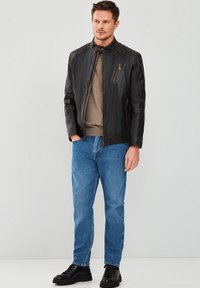 Chaqueta de cuero negra con acentos acolchados en los hombros, cuello corto y bolsillo en el pecho con cremallera. Combinada con una camisa gris, jeans azules y zapatos negros.