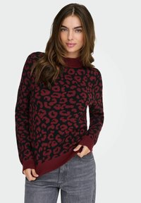 Pull en imprimé léopard noir et bordeaux avec un col et des poignets côtelés, doté d'une coupe décontractée et d'une texture douce. Associé à un jean gris.
