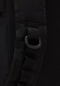 Sac à dos noir en tissu avec panneau en mesh, présentant des bretelles ajustables, un anneau en D en plastique et des coutures détaillées le long des bords.