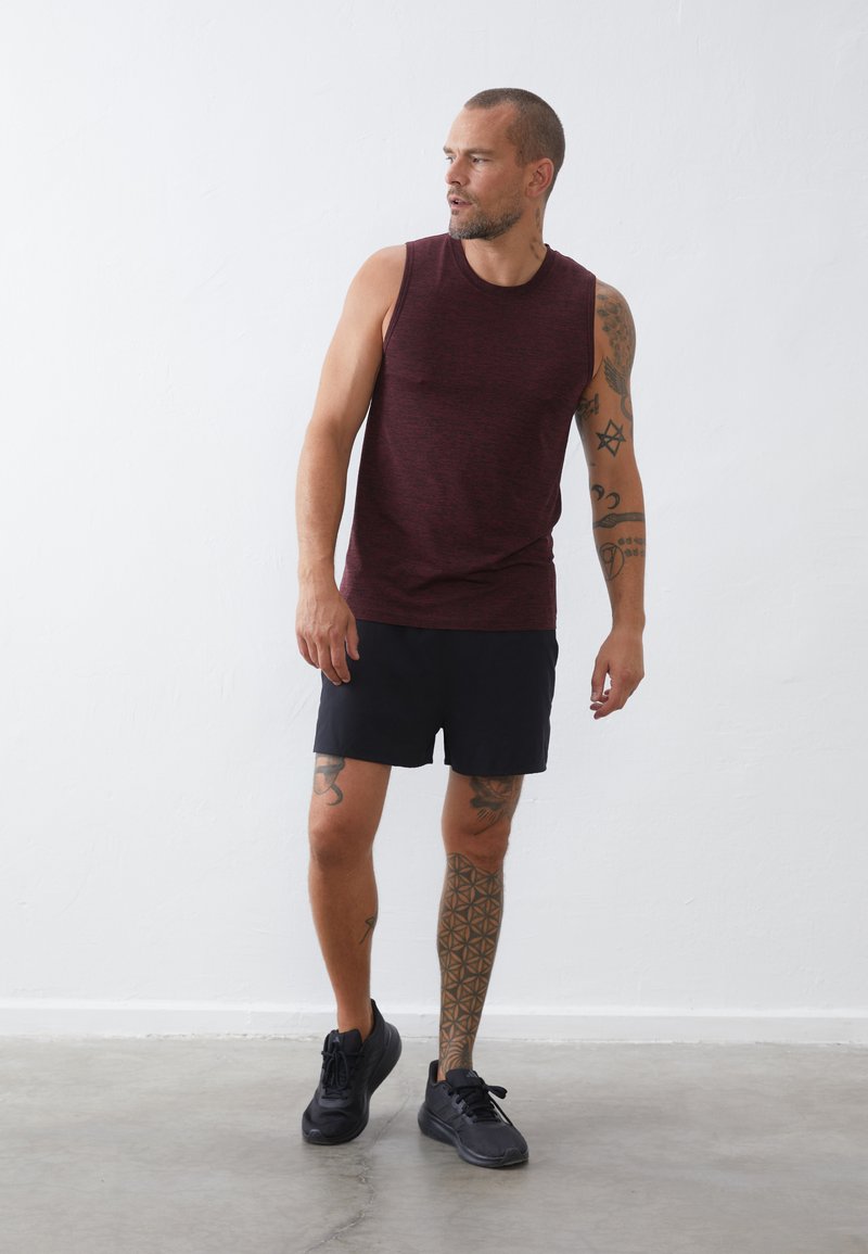 Μαρόν αμάνικο τοπ, μαύρες βερμούδες και μαύρα sneakers; έχει τατουάζ στο χέρι και το πόδι; στέκεται σε λείο γκρι πάτωμα με λευκό τοίχο στο φόντο.