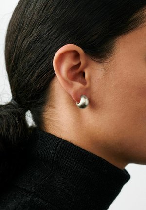Lähikuva henkilön korvasta, jossa on pieni hopeinen, puolikuun muotoinen korvakoru. Henkilöllä on tummat hiukset sidottuna ja musta pooloneule.