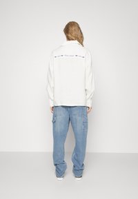 White linen shirt with long sleeves, back embroidered with blue arrows and text, paired with loose blue denim cargo pants.