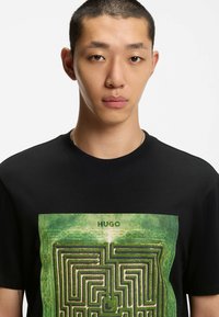 Zwarte T-shirt met een groene doolhofafbeelding op de voorkant en de tekst "HUGO" erboven. De stof lijkt glad en het shirt heeft een ronde hals.