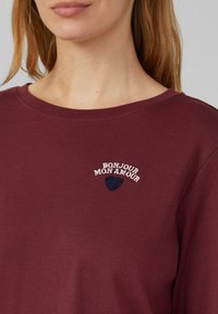 Burgunderfarbenes Langarmhemd mit weiß gesticktem Text "BONJOUR MON AMOUR" und einem dunkelblauen Herzdesign auf der Brust. Aus weichem Baumwollmaterial.