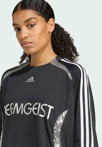 Čierna športová dres s striebornými flitrami, vybavený okrúhlym výstrihom, dlhými rukávmi a bielymi pruhmi Adidas pozdĺž ramien.