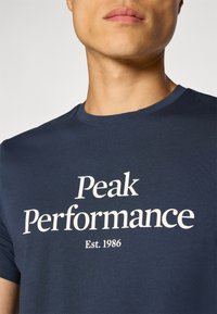 Camiseta de algodón azul marino con cuello redondo, que presenta un gran texto blanco que dice "Peak Performance" y "Est. 1986" debajo.