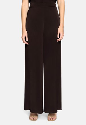 Kvinde iført højttaljede, vidde-sorte bukser kombineret med beige sandaler med åben tå, stående mod en ensfarvet hvid baggrund.