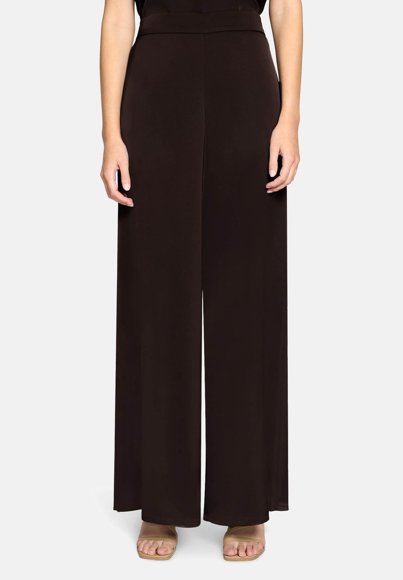 Femme portant un pantalon noir taille haute à jambes larges, associé à des sandales beiges à bout ouvert, debout devant un fond blanc uni.