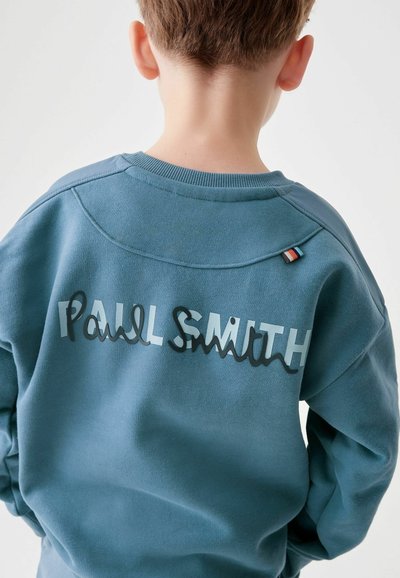 Sinine spordipusa, millel on tekstuuriga, ülekaalus sobivus. Omab "Paul Smith" logo kihilise musta ja helesinise letteringuga. Lisatud aktsent-silt.