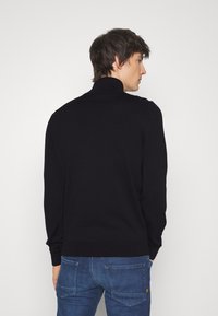 Schwarzer Strickpullover mit hohem Kragen, gerippten Bündchen und Saum, ergänzt durch eine glatte Textur, getragen mit blauen Denim-Jeans.