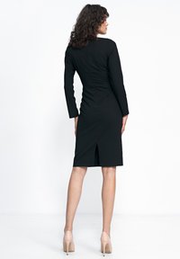 Czarna dopasowana sukienka z długimi rękawami, teksturowanym szwem z tyłu, sięgająca do kolan, z rozcięciem z tyłu na środku. Noszona w połączeniu z cielistymi szpilkami.