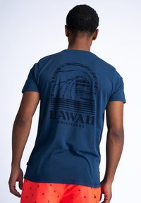Marineblauwe katoenen T-shirt met een golfafbeelding en de tekst "HAWAII WAVERIDERS" in zwart op de achterkant. Korte mouwen, ronde hals.