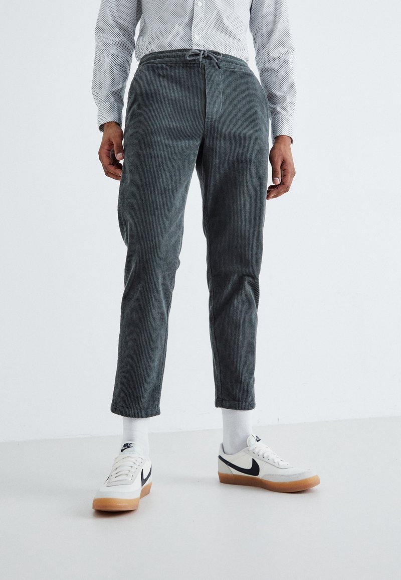Un homme porte un pantalon en velours côtelé gris, des chaussettes blanches, des baskets blanches Nike avec le swoosh noir, et une chemise à manches longues légère à motifs, debout sur un sol blanc.