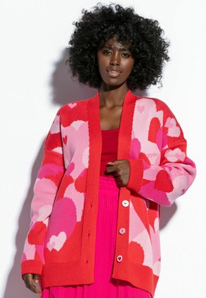 Czerwony i różowy kardigan z wzorem serc, luźny fason, teksturowany splot, z dużymi guzikami i ściągaczami na mankietach, w połączeniu z czerwonym topem.