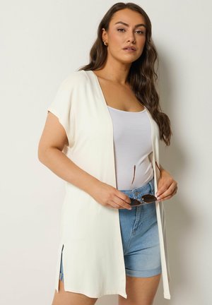 Donna con capelli lunghi e scuri che indossa un cardigan bianco senza maniche sopra una canottiera bianca e pantaloncini di jeans, tiene un paio di occhiali da sole con entrambe le mani.